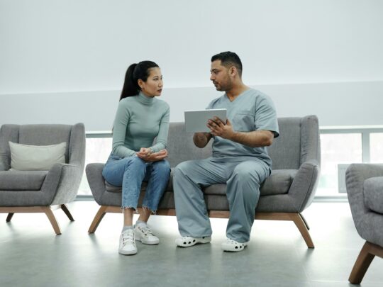 nurse in gray scrubs speaking with a patient on a couch